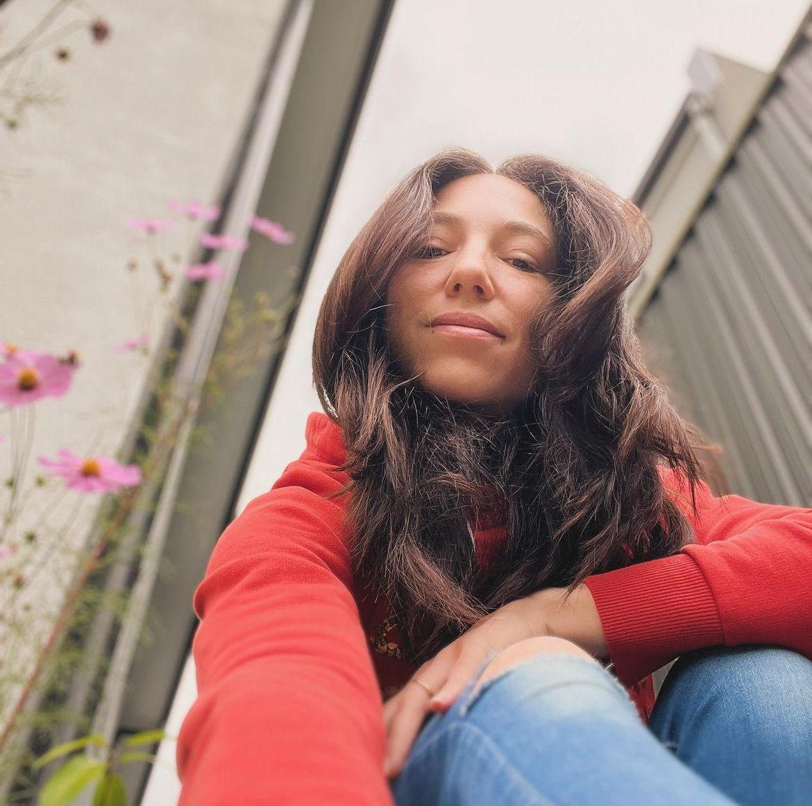 Person wearing a red sweatshirt and blue jeans, sitting outdoors near a flowering plant with pink blossoms.