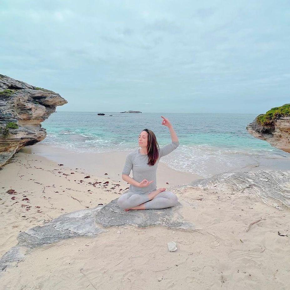 Person practicing yoga on a sandy beach with rocky cliffs and calm blue ocean in the background.