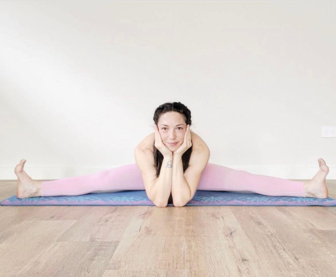 Person wearing pink leggings performing a wide-legged split on a blue yoga mat on a wooden floor.