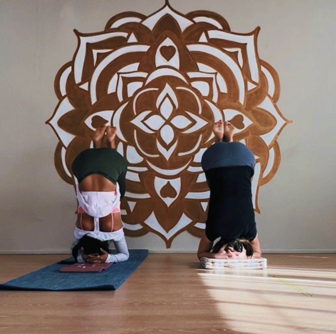 Two people performing headstands on yoga mats against a wall with a decorative mandala design.