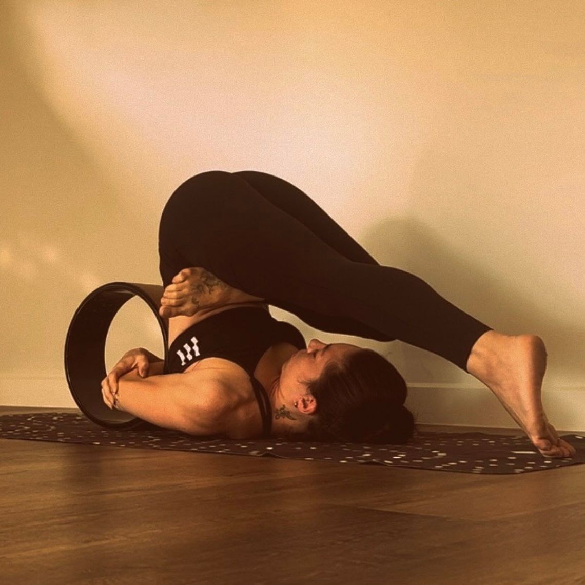 Person practicing an advanced yoga pose using a yoga wheel on a mat indoors.