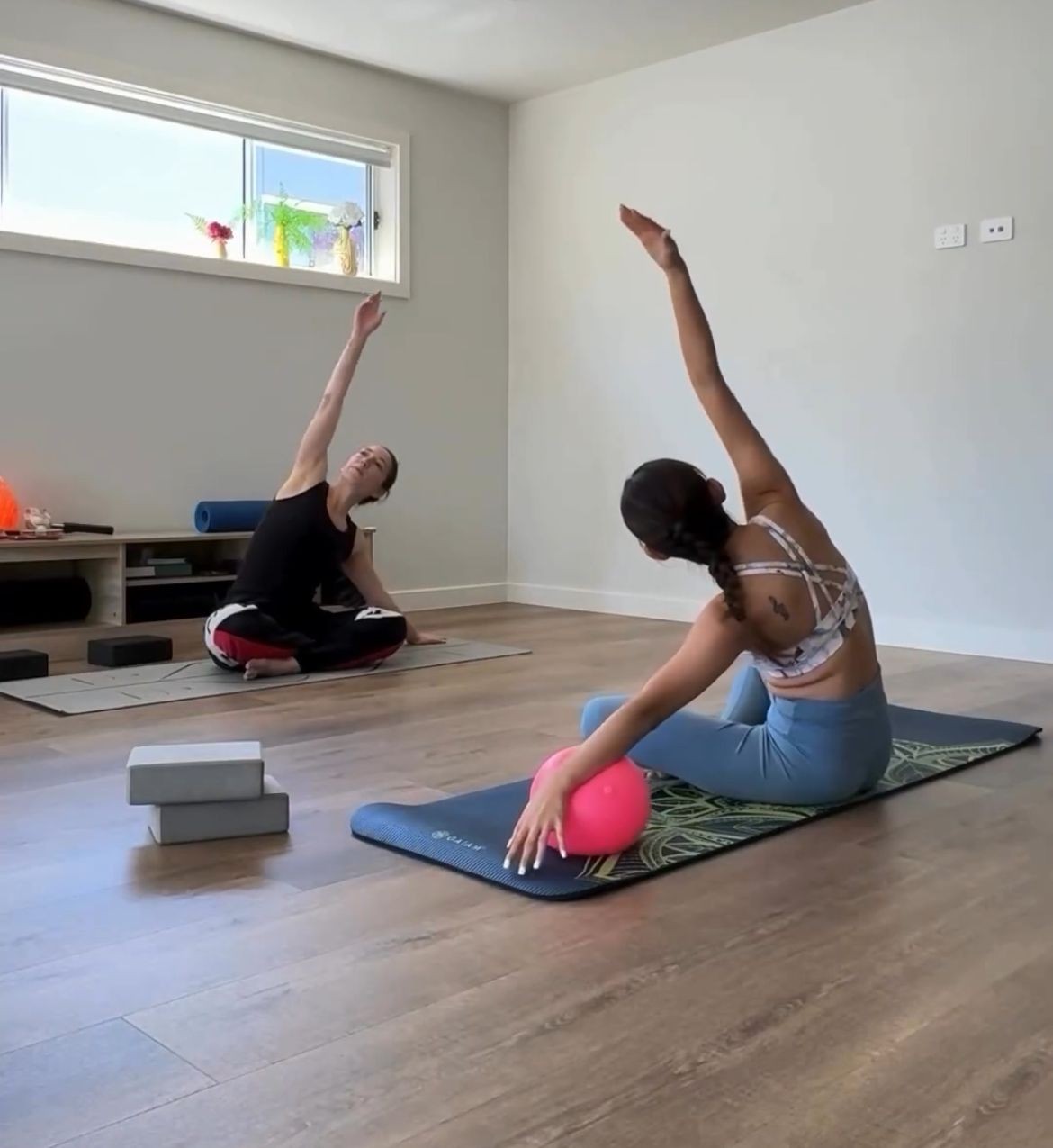 Two people doing yoga stretches on mats with a pink ball in a bright room.