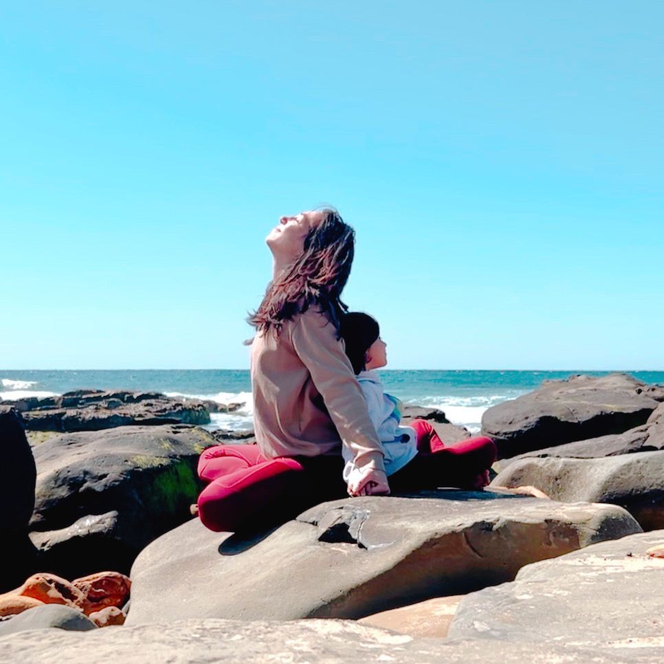 Woman and child sitting on rocks by the sea under a clear blue sky, enjoying the scenery.